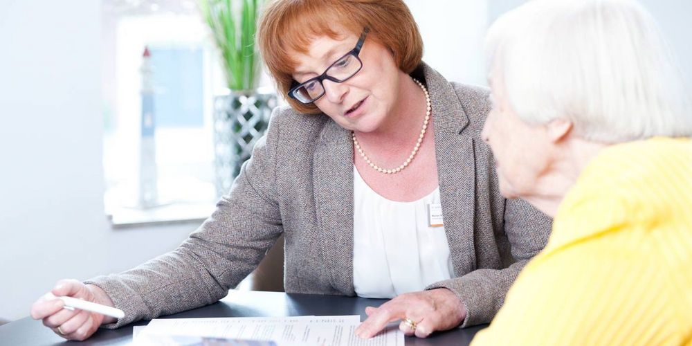Eine Frau sitzt mit einer Seniorin an einem Tisch und berät diese. 