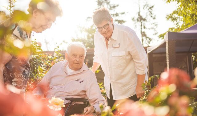 Eine Seniorin und ein Senior im Rollstuhl sind mit einer Frau im Garten und betrachten die Pflanzen.