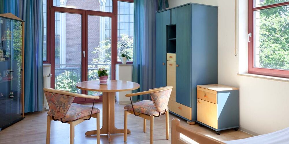 Zimmer im Haus Theresienheim mit einem Bett, zwei Stühlen, einem Tisch und einem großen sowie kleineren Schränken. 