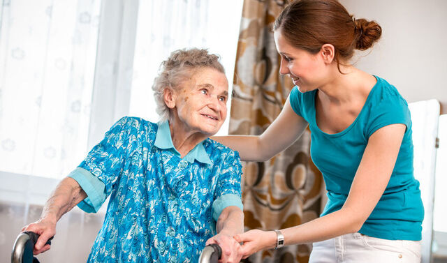 Eine Seniorin geht an einer Gehhilfe und bekommt hierbei Unterstützung von einer jungen Pflegerin. Die beiden schauen sich an.