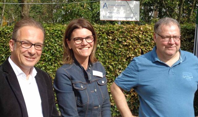 Eine Frau steht in der Mitte zwischen zwei Herren. Alle drei stehen draußen im Freien. 