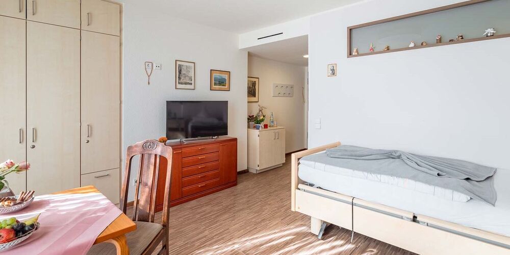 Zimmer im Johannes von Gott Haus mit einem Einzelbett, Tisch, Stühlen und einer Kommode mit Fernseher. 