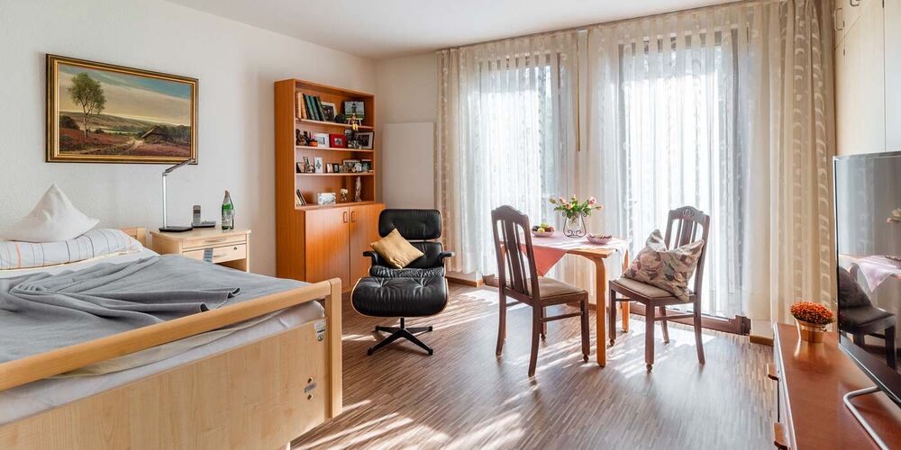 Zimmer im Johannes von Gott Haus. In diesem steht ein Bett, ein Sessel, ein Tisch mit zwei Stühlen ud ein Regal. Außerdem steht neben dem Tisch ein Nachttisch und vor dem Bett eine Kommode mit einem Fernseher. 