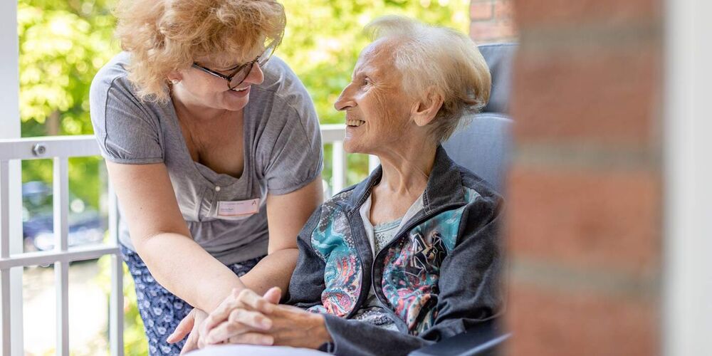 Eine Seniorin sitzt draußen auf einem Balkon. Eine Frau beugt sich zu ihr runter und beide schauen einander an. 
