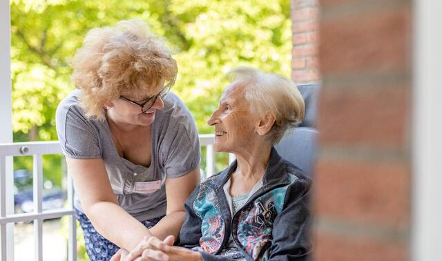 Eine Seniorin sitzt draußen auf einem Balkon. Eine Frau beugt sich zu ihr runter und beide schauen einander an.