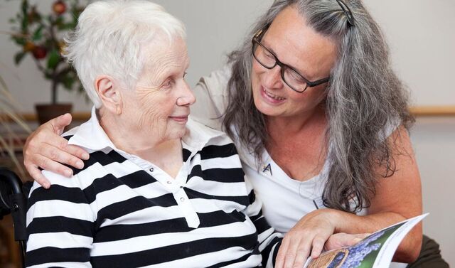 Eine Seniorin hält eine Zeitung in der Hand. Eine Frau sitzt neben ihr und hat die Hand auf der Zeitschrift. Die beiden Frauen schauen einander an. 