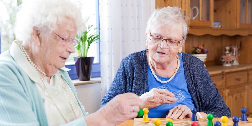 Zwei Seniorinnen sitzen an einem Tisch und spielen Mensch ärgere dich nicht. 