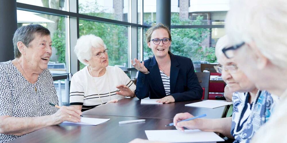 Einige Seniorinnen sitzen mit einer Frau an einem Tisch. Sie unterhalten sich angeregt und haben Papiere vor sich auf dem Tisch liegen.