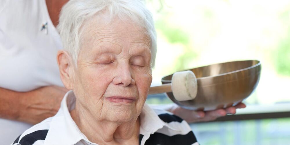 Eine Seniorin sitzt mit geschlossenen Augen auf einem Stuhl. Eine Dame steht hinter ihr und schlägt neben ihm mit einem Stab an eine Klangschale.