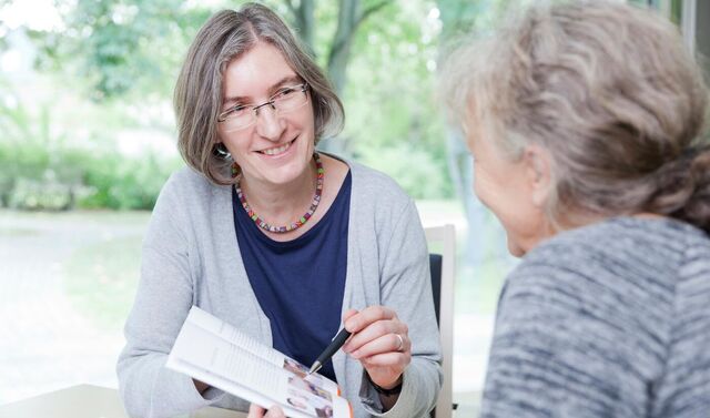Zwei Damen sitzen an einem Tisch in einer Beratungssituation. Die eine der beiden Damen hat einen Flyer in der Hand und zeigt etwas mit einem Stift auf diesem. 