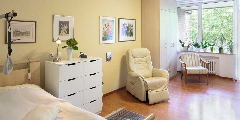 Zimmer der Seniorenhilfe des Haus Maria Hilf. In diesem steht ein Bett, ein Sessel sowie eine Kommode und ein Schrank. Es hängen einige Bilder an den Wänden und es sind Pflanzen im Zimmer. 